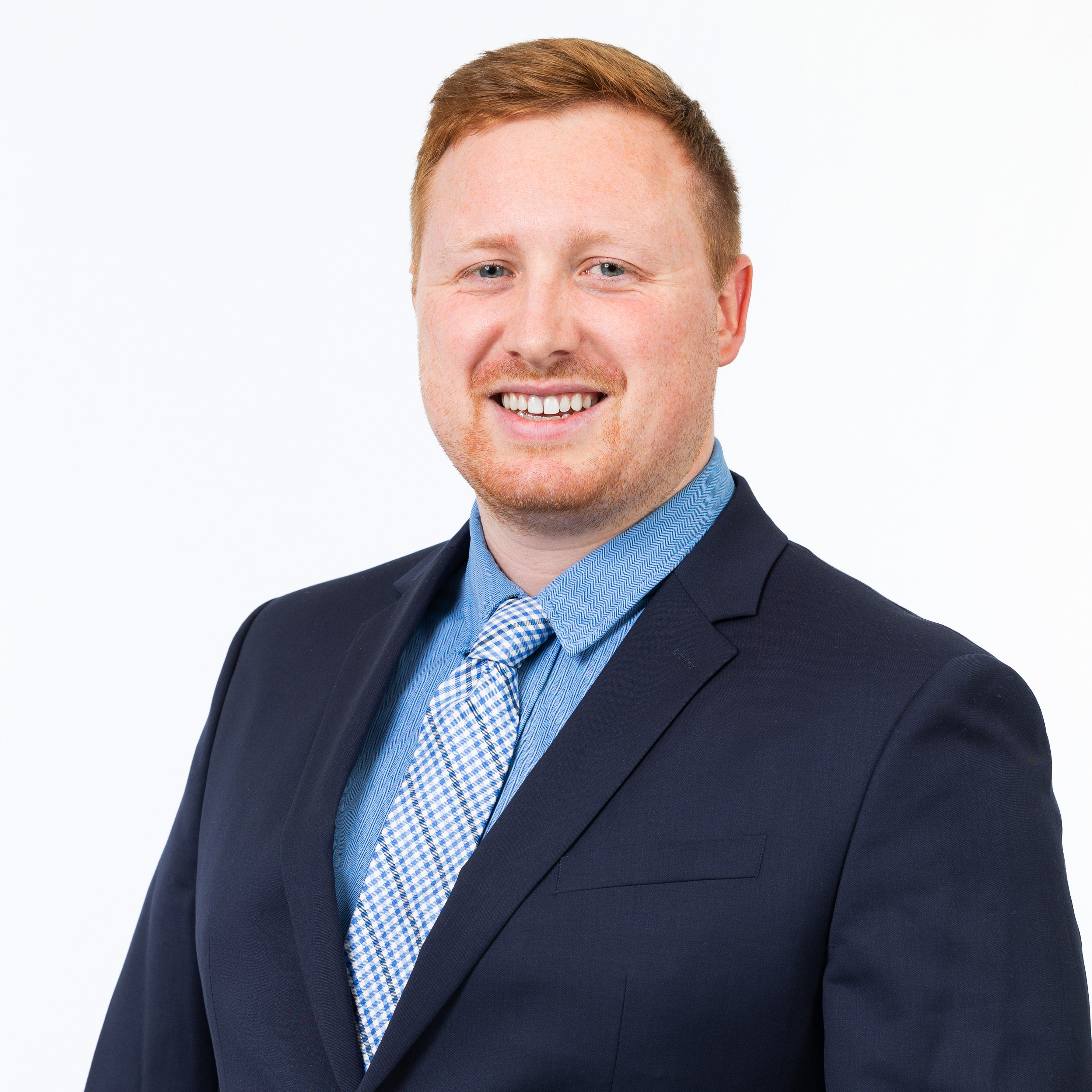 Headshot of a School of Social Work administration member with short red hair and light stubble, smiling, wearing a navy suit jacket, blue shirt, and patterned tie against a light gray background.
